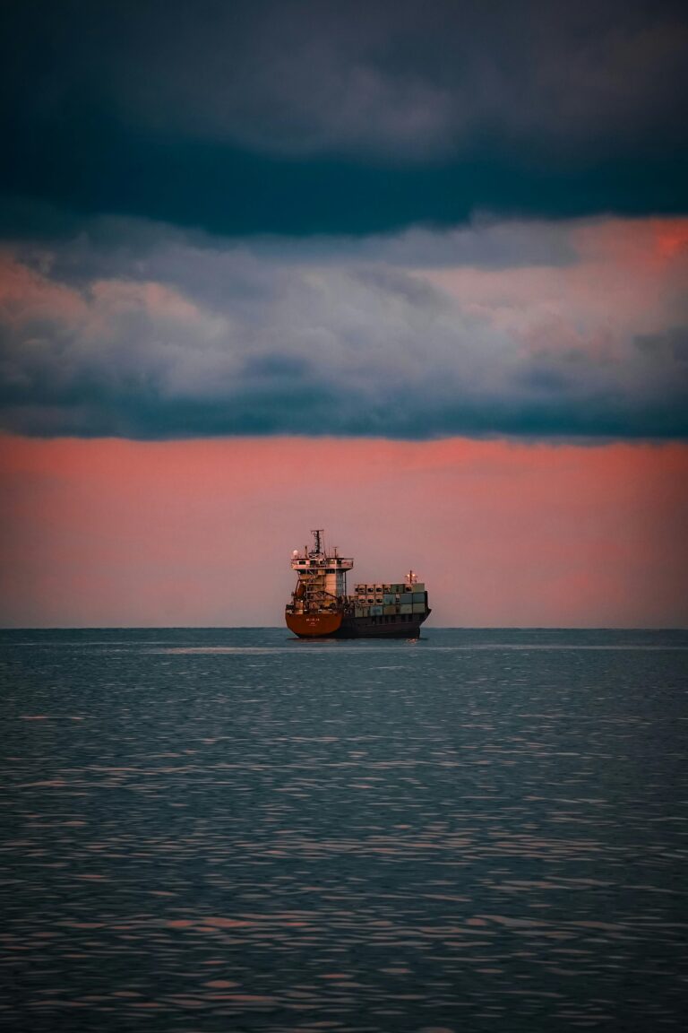 A solitary cargo ship sails under a dramatic sky with sunset hues over the calm sea.