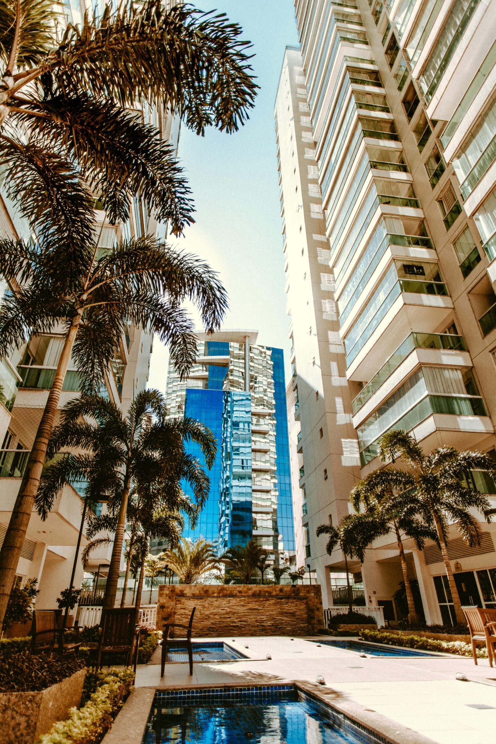 A modern cityscape with tall skyscrapers and palm trees surrounding a serene pool.
